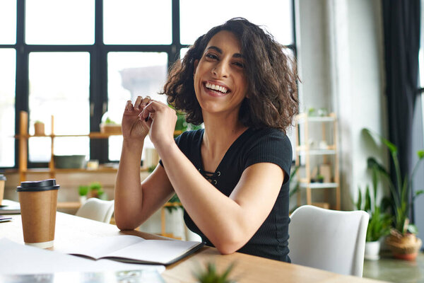 happiness and inspiration, overjoyed multiracial coach sitting with pen and looking at camera near notebook and coffee to go in women interest club, social activism and dedication concept