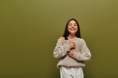 Cheerful preteen girl in stylish knitted sweater and casual outfit touching chest while looking at camera and standing isolated on green, contemporary fashion for preteen concept  clipart