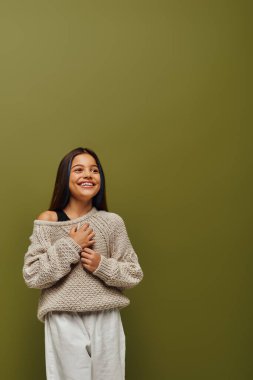 Joyful brunette preteen girl in knitted sweater and casual outfit touching chest while looking away and standing on green background, contemporary fashion for preteen concept clipart