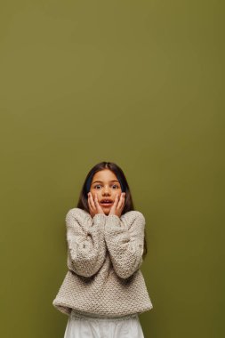 Şok olmuş esmer, saçı gündelik ve şık örgü süveter giyen, yanaklarına dokunan ve gençlik konsepti için çağdaş moda olan yeşil kameraya bakan genç kız.