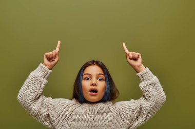 Shocked preteen girl with colored hair wearing stylish knitted sweater while pointing with fingers and looking at camera isolated on green, fashion-forward preteen with sense of style clipart