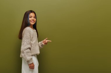 Gülümseyen esmer, boyalı saçlı, modaya uygun örgü süveter giyen ve yeşil arka plandaki kameraya bakarken parmağıyla işaret eden, modaya uygun, modaya uygun, genç bir kız.