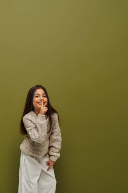 Süveter örgülü saçlarıyla neşeli ve şık bir genç kız gizli jestler yapıyor ve yeşil arka planda dururken kameraya bakıyor, modern ve modaya uygun.