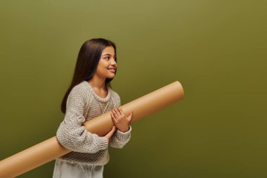 Smiling brunette preadolescent girl in trendy knitted sweater and outfit holding rolled paper and looking away isolated on green, stylish girl in cozy fall attire concept clipart