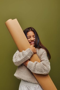 Pleased and stylish preadolescent girl with dyed hair wearing knitted sweater while hugging rolled paper and standing isolated on green, girl radiating autumn vibes concept clipart
