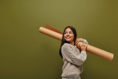 Positive and trendy preadolescent girl with dyed hair wearing autumn knitted sweater and looking away while holding rolled paper isolated on green, girl radiating autumn vibes concept clipart