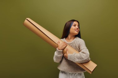 Smiling preadolescent girl with dyed hair wearing autumn outfit and knitted sweater while looking away and holding rolled paper isolated on green, girl radiating autumn vibes clipart