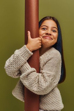 Portrait of excited and smiling trendy preteen girl in knitted sweater hugging rolled paper and looking at camera while standing isolated on green, girl radiating autumn vibes concept clipart