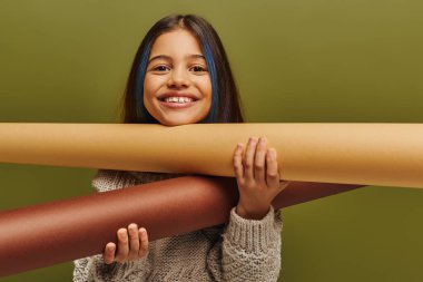 Saçları boyalı, kalın örgü süveter giyen ve elinde sarılı kağıtlarla genç kızlar için yeşil, sonbahar modasında tek başına duran pozitif ergenlik öncesi çocuk portresi.