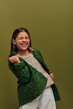 Excited and stylish preadolescent girl with dyed hair showing yes gesture while posing in autumn jacket and knitted sweater isolated on green, modern fall fashion for preteens concept clipart