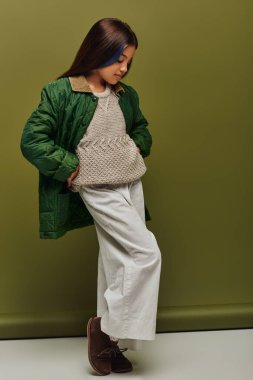 Sonbahar ceketiyle poz veren ve yeşil arka planda dururken örgü ören esmer, modaya uygun, genç kızlar için modern moda.