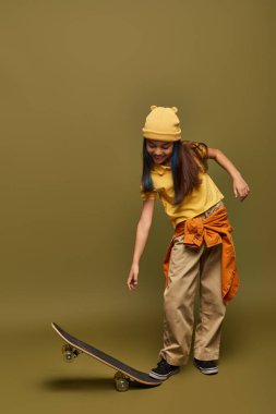 Boyalı saçlı, sarı şapkalı ve şehir kıyafetli neşeli ergen çocuk haki rengi arka planda kaykaya bakarken modern kıyafet konseptinde şık bir kız.
