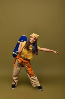 Full length of positive preteen girl with dyed hair wearing yellow hat and urban outfit while holding skateboard and standing on khaki background, stylish girl in modern outfit concept clipart