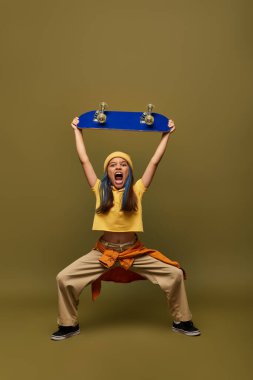 Mad and stylish preteen girl with dyed hair wearing stylish yellow hat and outfit and holding skateboard and standing on khaki background, girl with cool street style look clipart