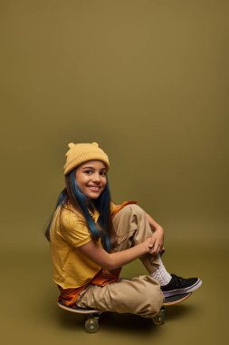 Smiling preteen and stylish child with dyed hair wearing urban outfit and hat looking at camera while sitting on skateboard on khaki background, girl with cool street style look clipart