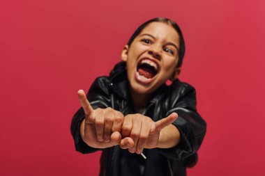 Blurred and mad preadolescent girl in stylish leather jacket showing rock gesture at camera while standing and posing isolated on red, girl with cool and contemporary look clipart