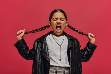 Mad and fashionable preadolescent girl screaming and touching hairstyle while posing in leather jacket and white t-shirt and standing isolated on red, girl with cool and contemporary look clipart