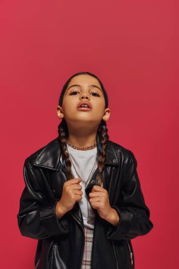 Portrait of fashionable preteen girl touching hair while posing in modern leather jacket and looking at camera and standing isolated on red, girl with cool and contemporary look clipart