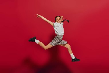 Full length of pleased and stylish preteen girl with hairstyle wearing t-shirt and plaid skirt while jumping and having fun on red background, hairstyle and trendy accessories concept clipart