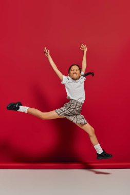 Excited and cheerful preteen girl in white t-shirt and stylish checkered skirt jumping and having fun while posing on red background, hairstyle and trendy accessories concept clipart