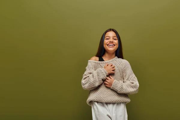 stock image Cheerful preteen girl in stylish knitted sweater and casual outfit touching chest while looking at camera and standing isolated on green, contemporary fashion for preteen concept 