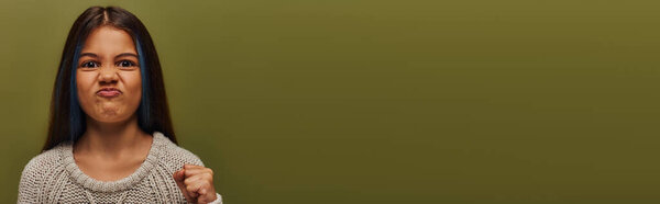 Portrait of angry preteen child with dyed hair wearing knitted sweater and looking at camera while holding hand in fist isolated on green, stylish girl in cozy fall attire, banner with copy space