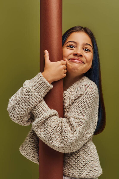 Portrait of excited and smiling trendy preteen girl in knitted sweater hugging rolled paper and looking at camera while standing isolated on green, girl radiating autumn vibes concept