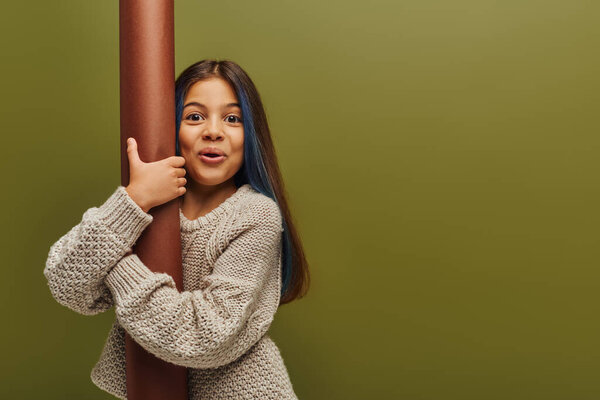 Excited and trendy preadolescent girl with dyed hair wearing knitted sweater looking at camera while hugging rolled paper and standing isolated on green, girl radiating autumn vibes