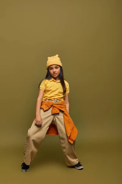 stock image Full length of confident preadolescent girl in urban outfit and yellow hat posing and looking at camera while standing on khaki background, stylish girl in modern outfit concept