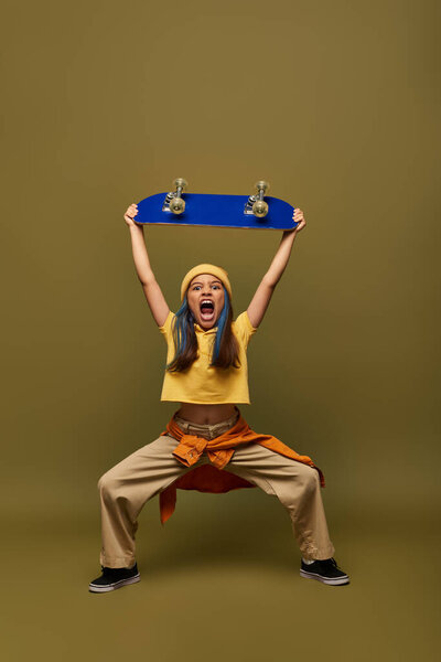 Mad and stylish preteen girl with dyed hair wearing stylish yellow hat and outfit and holding skateboard and standing on khaki background, girl with cool street style look