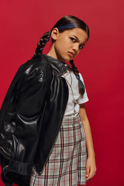 Displeased and stylish preteen girl with modern hairstyle wearing leather jacket and plaid skirt and looking at camera while standing isolated on red, stylish preteen outfit concept
