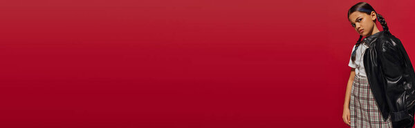 Недовольная девушка-подросток с прической в кожаной куртке и клетчатой юбке, глядя в камеру и стоя изолированно на красной, стильной концепции детского наряда, баннер 