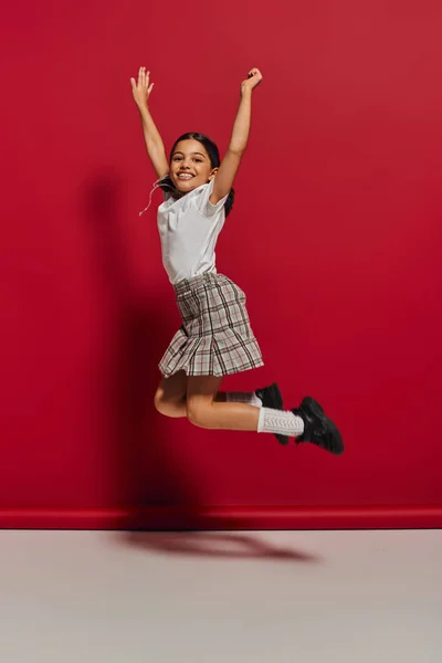 stock image Positive and trendy brunette preteen girl in t-shirt and checkered skirt jumping while having fun and looking at camera on red background, hairstyle and trendy accessories concept