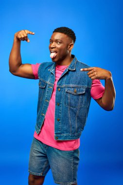 Stylish young african american man with modern hairstyle sticking out tongue and pointing with fingers while posing in summer outfit isolated on blue, trendy man showing summer style clipart