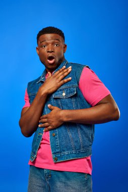 Shocked young african american man with modern hairstyle wearing denim vest and bright t-shirt while looking at camera and standing isolated on blue, trendy man showing summer style clipart