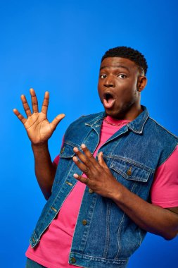 Astonished young african american man with modern hairstyle in bright pink t-shirt and denim vest looking at camera while standing isolated on blue, trendy man showing summer style clipart