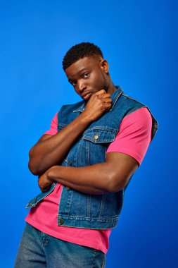 Confident young african american man with modern hairstyle wearing denim vest and bright pink t-shirt and looking at camera while standing isolated on blue, trendy man showing summer style clipart