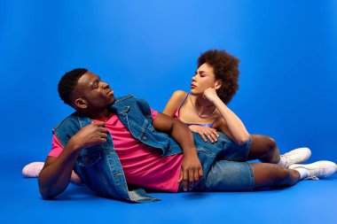 Düşünceli genç Afro-Amerikan en iyi arkadaşları, parlak ve şık yaz kıyafetleri içinde mavi arka planda poz verirken birbirlerine bakıyorlar, moda kankaları güven yayıyor, arkadaşlık kuruyorlar.