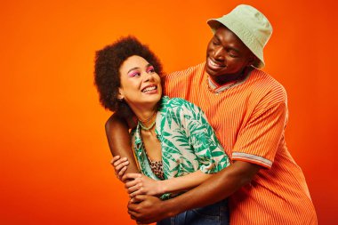 Panama şapkalı Afro-Amerikan genç adam cesur makyajlı modaya uygun en iyi arkadaşı kucaklıyor ve birbirlerine turuncu üzerinde izole bir şekilde bakıyor, arkadaşlar bireysel bir tarz sergiliyor.