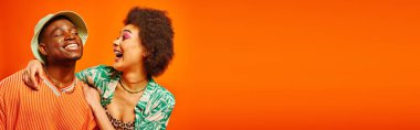 Excited young african american woman with bold makeup hugging cheerful best friend in summer outfit and panama hat isolated on orange, friends showcasing individual style, banner  clipart