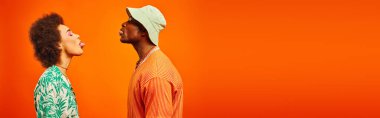 Side view of young and fashionable african american best friends in summer outfits sticking out tongues while standing together isolated on orange, banner, friends in trendy clothes clipart