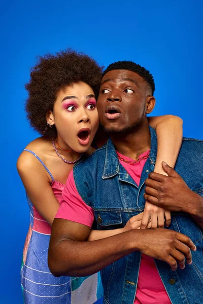 stock image Shocked young african american woman with bold makeup wearing sundress and hugging stylish best friend in denim vest while standing isolated on blue, fashionable friends in trendy clothes