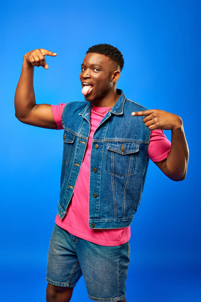 Stylish young african american man with modern hairstyle sticking out tongue and pointing with fingers while posing in summer outfit isolated on blue, trendy man showing summer style