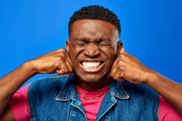 Angry young african american man with modern hairstyle wearing summer outfit and holding hands in fists while standing and posing isolated on blue, trendy man showing summer style