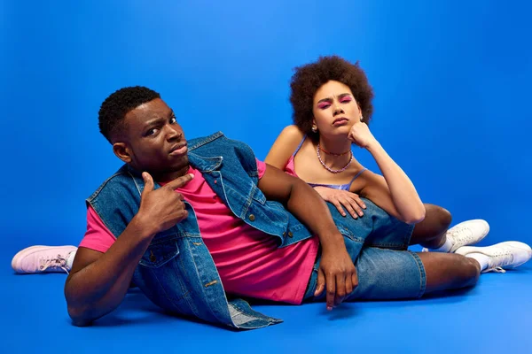 stock image Pensive young african american best friends in bright and trendy summer outfits looking at camera while lying together on blue background, fashionable besties radiating confidence, friendship