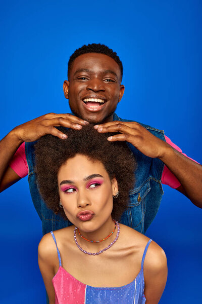 Smiling young african american man with modern hairstyle and summer outfit touching hair of best friend with bold makeup isolated on blue, fashionable besties radiating confidence, friendship