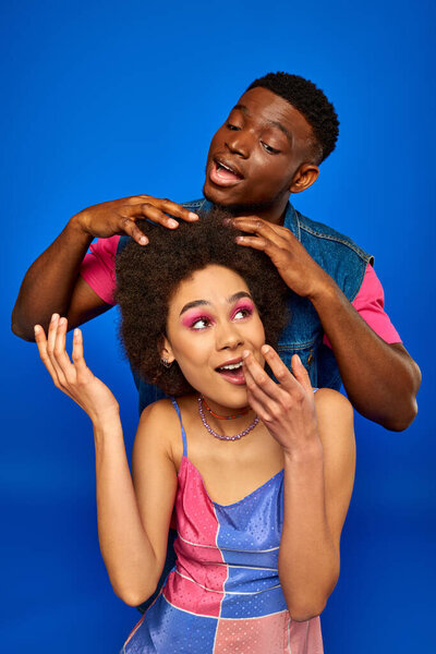 Young african american man with modern hairstyle touching hair of best friend in stylish summer sundress and standing isolated on blue, fashionable besties radiating confidence 