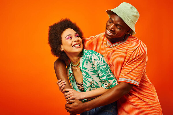 Приятно молодой африканский американец в панамской шляпе, обнимающий модного лучшего друга со смелым макияжем и смотрящий друг на друга изолированными от оранжевого, друзья демонстрирующие индивидуальный стиль