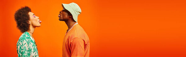Side view of young and fashionable african american best friends in summer outfits sticking out tongues while standing together isolated on orange, banner, friends in trendy clothes