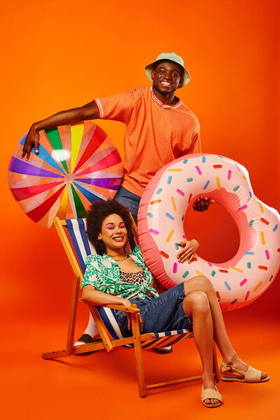 Full length of cheerful african american man in summer outfit holding pool ring and ball while standing near best friend on deck chair on orange background, fashion-forward friends, friendship